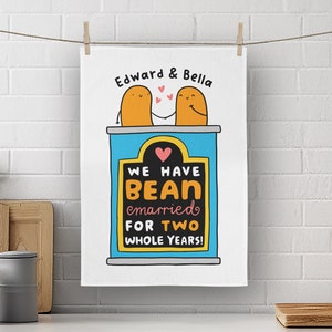 May include: A white kitchen towel with a blue and yellow illustration of two bean characters holding hands. The text reads "Edward & Bella We Have Bean Married For Two Whole Years!"