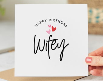 May include: White card with the text "Happy Birthday Wifey" in black script font. There are three small hearts above the text, one red and two pink.