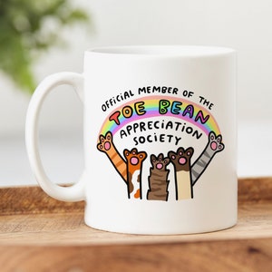 Puede incluir: Taza de cerámica blanca con un arcoíris y el texto "Official Member of the Toe Bean Appreciation Society". Tres gatos de dibujos animados con las patas extendidas están debajo del arcoíris.