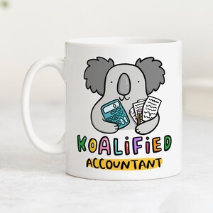 Op de afbeelding: Witte keramische mok met een cartoon koala die een rekenmachine, papieren en een potlood vasthoudt. De mok heeft de tekst "KOALIFIED ACCOUNTANT" in kleurrijke letters. Een leuk cadeau voor accountants.