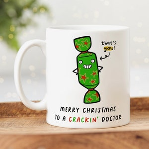 May include: White ceramic mug featuring a green and red Christmas cracker cartoon character. The text on the mug reads "that's you!" and "Merry Christmas to a Crackin' Doctor".