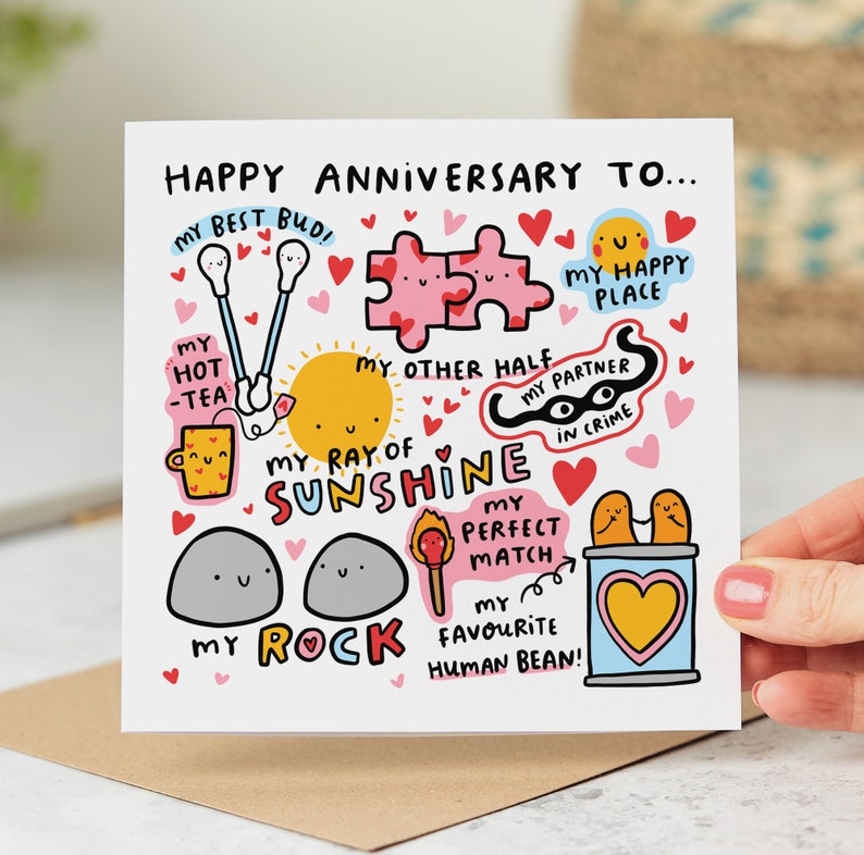 Op de afbeelding: Een witte kaart met een kleurrijke illustratie van harten, een zon, een theekopje, een puzzel, een rots, een lucifer en een blik. De tekst op de kaart luidt "Happy Anniversary To... My Best Bud, My Hot -Tea, My Other Half, My Ray of Sunshine, My Partner in Crime, My Perfect Match, My Rock, My Favorite Human Bean!"