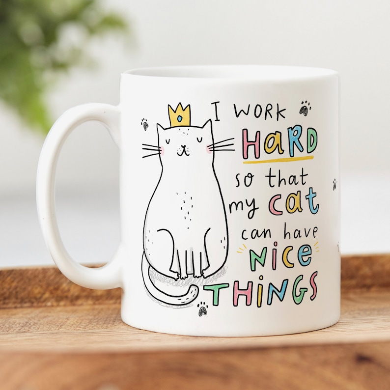 May include: White ceramic mug with a cartoon cat wearing a crown. The mug has the text "I work hard so that my cat can have nice things" in colorful letters.