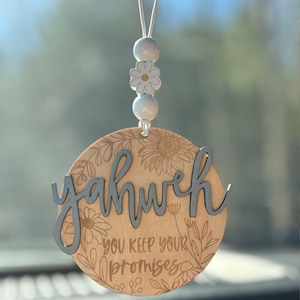 May include: A wooden car charm with the word "Yahweh" in light blue script. The charm is round with floral engravings and the text "You keep your promises." It hangs from a string with white beads and a daisy.