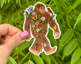 May include: A brown Bigfoot sticker with a variety of colorful flora and fauna. The sticker features a squirrel, butterfly, flowers, and a bird. The sticker is held in front of a background of green leaves.