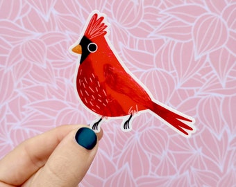 May include: A red cardinal bird sticker with a black beak and eye. The sticker is on a pink background with a white floral pattern.