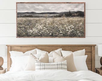 May include: A framed landscape painting depicting a field of wildflowers with mountains in the background. The artwork is displayed above a bed with white bedding and decorative pillows. The frame is brown.