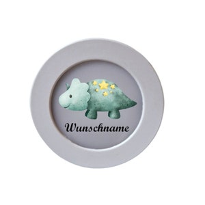 Könnte beinhalten: Ein rundes, grau gerahmtes Bild mit einer Aquarellillustration eines grünen Dinosauriers mit gelben Sternen. Der Text "Wunschname" steht in schwarzer Schrift unter dem Dinosaurier.