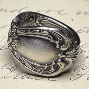 May include: A silver ring with an ornate, floral design. The ring features a detailed pattern of swirling leaves and flowers around the oval face. The metal has a polished, reflective surface, and the ring appears to be vintage or antique.