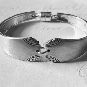 May include: A silver bracelet made from repurposed silverware. The bracelet features a magnetic clasp and decorative floral patterns on the ends. The design is a unique, handcrafted piece of jewelry.