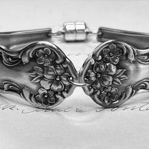 May include: A silver bracelet crafted from repurposed silverware. The bracelet features floral detailing on the spoon heads, connected by a magnetic clasp. The design showcases a vintage aesthetic, perfect for adding a unique touch to any outfit.