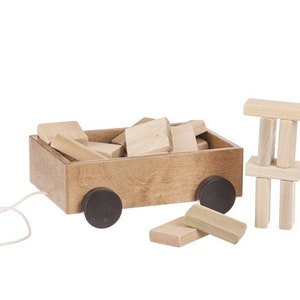 May include: A wooden toy wagon filled with wooden blocks. The wagon has black wheels and a rope attached to the front. There are also additional wooden blocks stacked on top of each other.