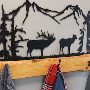 May include: A black metal wall-mounted coat rack featuring a mountain and forest silhouette design. The rack includes a natural wood base with three black metal hooks. Three articles of clothing hang from the hooks.
