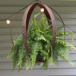 May include: A hanging planter with a rustic, weathered metal frame, holding a lush green fern. The circular design is suspended by a chain, with string lights adding a warm glow. A charming outdoor decor piece.