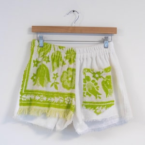 May include: White terry cloth shorts with a green floral and bird pattern. The shorts have an elastic waistband and a fringed hem. The shorts are hanging on a wooden hanger against a white background.