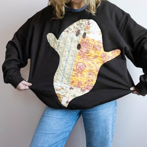 May include: A black long-sleeve sweatshirt featuring a large, quilted ghost-shaped appliqué on the front. The ghost is constructed from various patches of floral and patterned fabric in shades of yellow, cream, and orange, with two dark buttons for eyes.