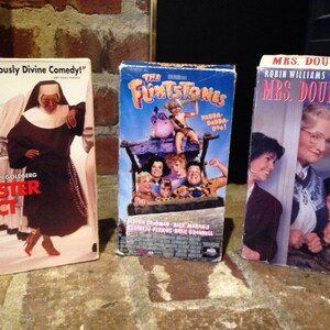 May include: Three VHS tapes, one with a blue background and the title "The Flintstones", one with a red background and the title "Sister Act" featuring Whoopi Goldberg, and one with a white background and the title "Mrs. Doubtfire" featuring Robin Williams and Sally Field.