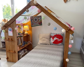 May include: A wooden house-shaped bed frame with a grey patterned bedspread. The bed is decorated with string lights and pillows. A shelf with toys and a lamp is visible inside the house structure. A decorative wall hanging of leaves is on the wall.
