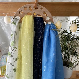 May include: A wooden moon-shaped hanger with four scarves hanging from it. The scarves are yellow with gold stars, navy blue with gold stars, and light blue with white stars. The hanger is decorated with stars and a crescent moon.