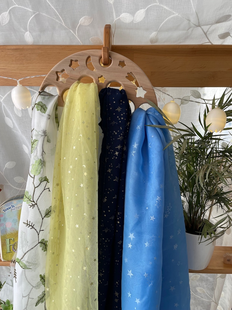 May include: A wooden moon-shaped hanger with four scarves hanging from it. The scarves are yellow with gold stars, navy blue with gold stars, and light blue with white stars. The hanger is decorated with stars and a crescent moon.