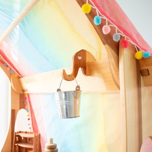 May include: A wooden playhouse with a rainbow-colored fabric canopy and a small metal bucket hanging from a wooden elephant hook. The playhouse has a wooden shelf with a wooden stacking toy and a small wooden house.