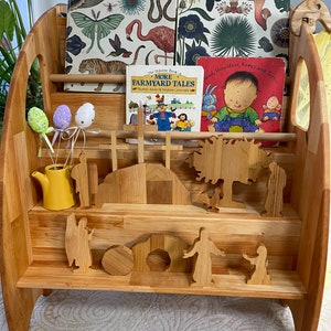 May include: A wooden bookshelf with three shelves, filled with children's books. The top shelf has two books with illustrations of animals and plants. The middle shelf has a book with illustrations of farm animals and a book with a child on the cover. The bottom shelf has wooden figurines of people and animals.