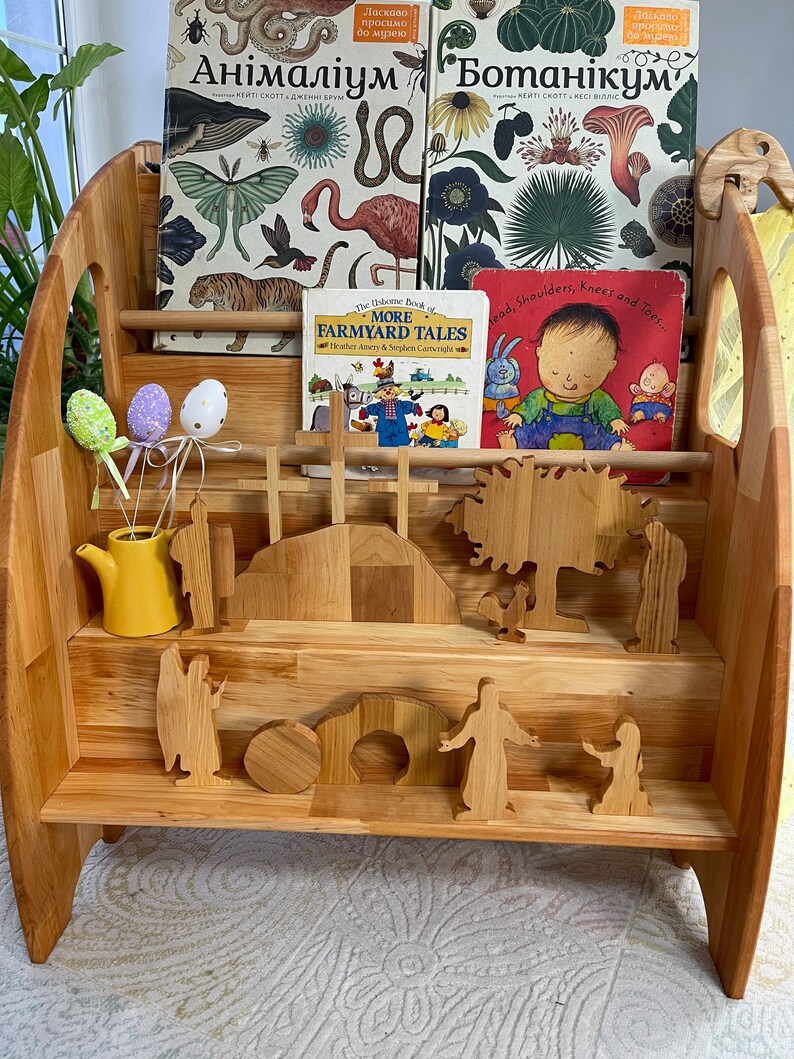May include: A wooden bookshelf with three shelves, filled with children's books. The top shelf has two books with illustrations of animals and plants. The middle shelf has a book with illustrations of farm animals and a book with a child on the cover. The bottom shelf has wooden figurines of people and animals.