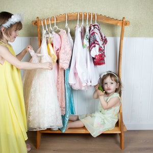 May include: Two young girls playing dress-up with a wooden clothing rack. The rack is filled with colorful dresses and shirts. One girl is wearing a yellow dress and the other is wearing a green dress.