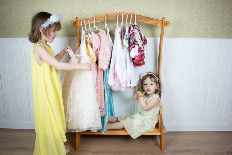 May include: Two young girls playing dress-up with a wooden clothing rack. The rack is filled with colorful dresses and shirts. One girl is wearing a yellow dress and the other is wearing a green dress.