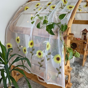 May include: White sheer curtain with embroidered yellow sunflowers. The curtain is hanging on a wooden frame with shelves.