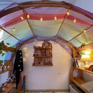 May include: A wooden play structure with a rainbow fabric canopy and string lights. The structure has shelves with toys and books. A small wooden dollhouse is on the wall.