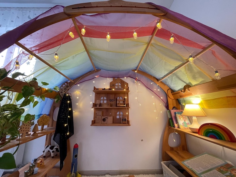 May include: A wooden play structure with a rainbow fabric canopy and string lights. The structure has shelves with toys and books. A small wooden dollhouse is on the wall.