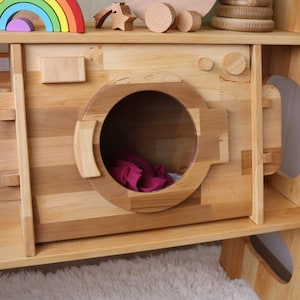 May include: A wooden toy washing machine with a large, round opening and a pink cloth inside. The washing machine is part of a larger wooden playset with shelves and other features.