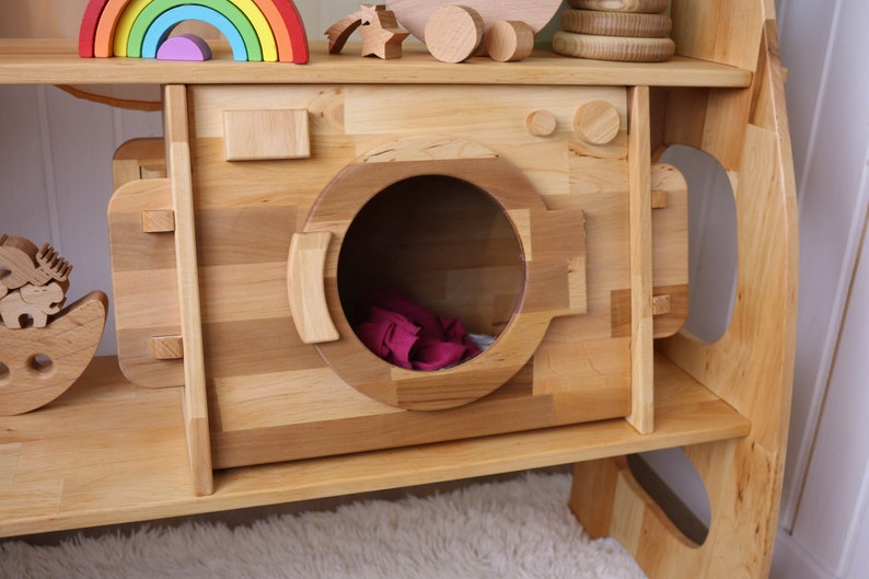 May include: A wooden toy washing machine with a large, round opening and a pink cloth inside. The washing machine is part of a larger wooden playset with shelves and other features.