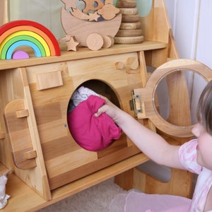 May include: A wooden toy washing machine with a pink fabric item inside. A child is opening the door of the washing machine.