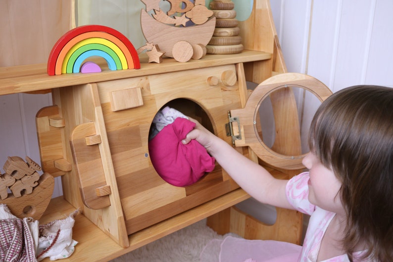 May include: A wooden toy washing machine with a pink fabric item inside. A child is opening the door of the washing machine.