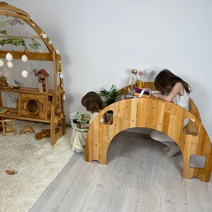 May include: Two children playing in a playroom with wooden play structures. One child is sitting at a wooden table and drawing, while the other child is sitting on the floor looking at a book. The playroom has a light wood floor and a white wall.
