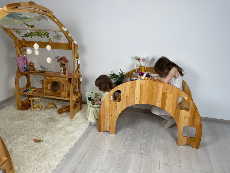 May include: Two children playing in a playroom with wooden play structures. One child is sitting at a wooden table and drawing, while the other child is sitting on the floor looking at a book. The playroom has a light wood floor and a white wall.