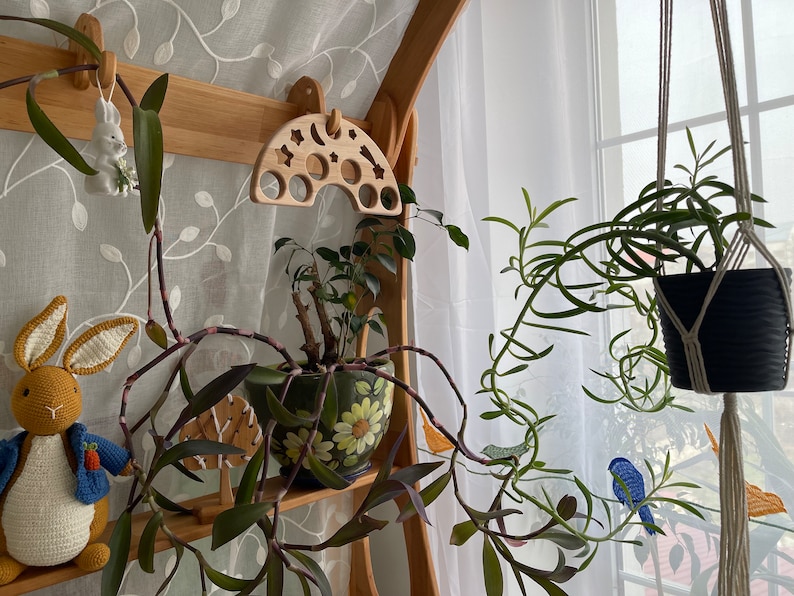 May include: A wooden rainbow-shaped toy with holes for stacking, hanging from a shelf with a white porcelain bunny figurine. A potted plant with green leaves and a hanging plant with green leaves in a black pot are also visible.