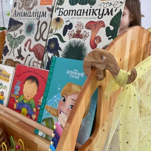 May include: A wooden bookshelf with a yellow tulle curtain and a variety of children's books, including "More Farm Yard Tales", "Head, Shoulders, Knees and Toes", and "Крижане Серце".