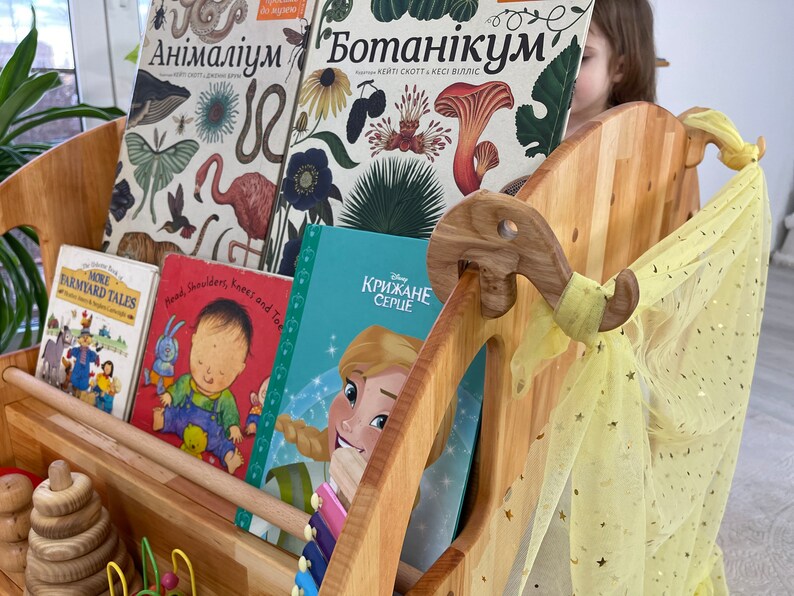 May include: A wooden bookshelf with a yellow tulle curtain and a variety of children's books, including "More Farm Yard Tales", "Head, Shoulders, Knees and Toes", and "Крижане Серце".
