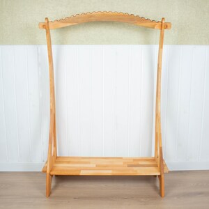 K&ouml;nnte beinhalten: Ein Kleiderst&auml;nder aus Holz mit einer gebogenen Oberseite und einem Regal. Der St&auml;nder ist aus Naturholz gefertigt und hat ein schlichtes, rustikales Design.