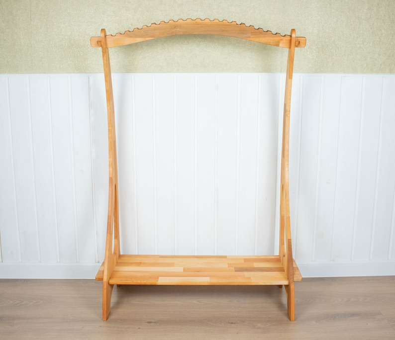 K&ouml;nnte beinhalten: Ein Kleiderst&auml;nder aus Holz mit einer gebogenen Oberseite und einem Regal. Der St&auml;nder ist aus Naturholz gefertigt und hat ein schlichtes, rustikales Design.