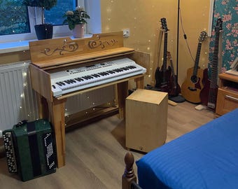 Könnte beinhalten: Eine weiße digitale Tastatur mit einem Holzkasten und verzierten Schnitzereien. Die Tastatur steht auf einem Ständer und im Hintergrund befinden sich drei Gitarren und ein Lautsprecher.