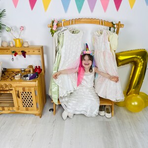K&ouml;nnte beinhalten: Ein junges M&auml;dchen in einem wei&szlig;en Kleid und einem Einhorn-Stirnband sitzt auf einem Holzhocker vor einem Kleiderst&auml;nder. Sie ist umgeben von bunten Dekorationen, darunter ein goldener Ballon mit der Nummer sieben und gelbe Ballons.