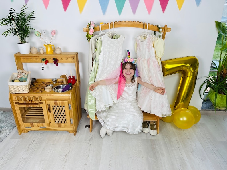 K&ouml;nnte beinhalten: Ein junges M&auml;dchen in einem wei&szlig;en Kleid und einem Einhorn-Stirnband sitzt auf einem Holzhocker vor einem Kleiderst&auml;nder. Sie ist umgeben von bunten Dekorationen, darunter ein goldener Ballon mit der Nummer sieben und gelbe Ballons.
