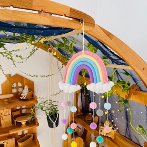 May include: A rainbow-coloured yarn rainbow mobile with white pom-poms hanging from it. The mobile is hanging from a wooden structure with a blue fabric canopy. The mobile is decorated with white clouds and colourful pom-poms.