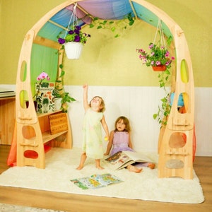 May include: Two children play in a wooden playhouse with a rainbow fabric canopy. The playhouse has a built-in bench and shelves. There are hanging plants and a white rug on the floor.
