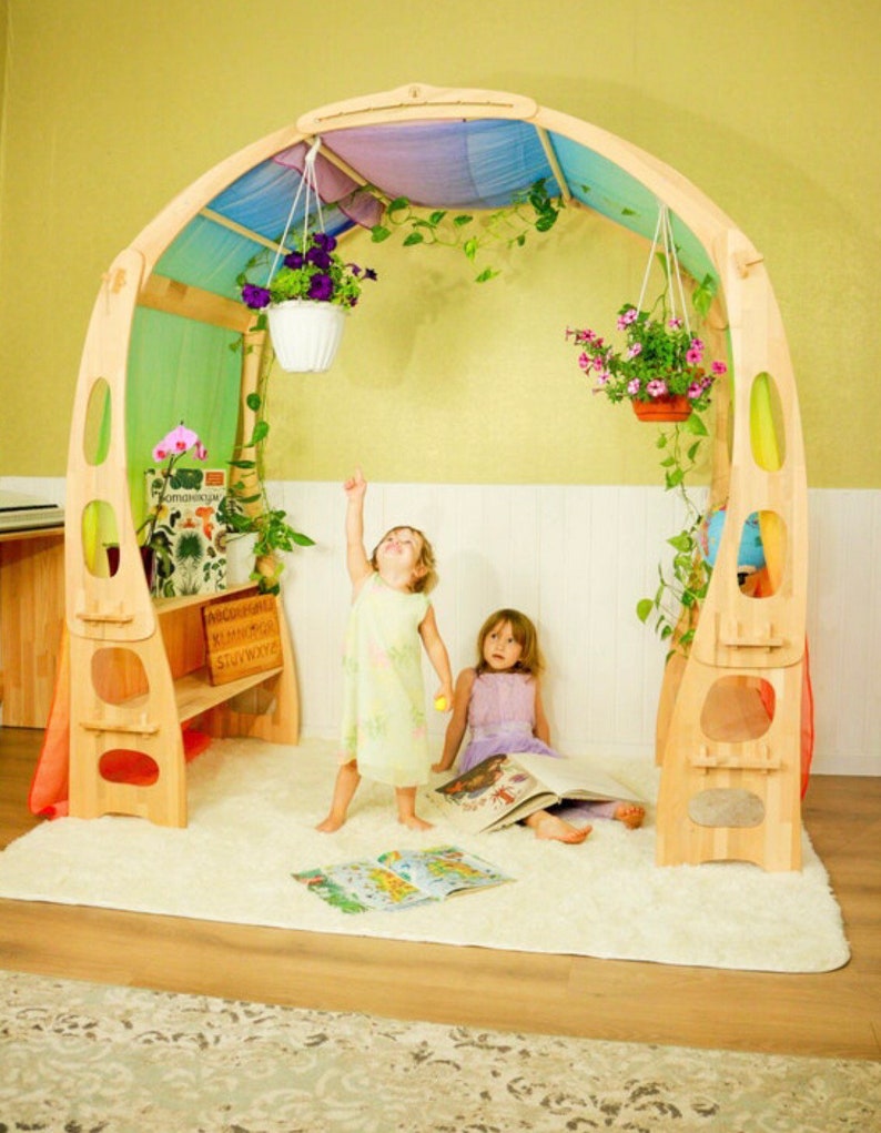 May include: Two children play in a wooden playhouse with a rainbow fabric canopy. The playhouse has a built-in bench and shelves. There are hanging plants and a white rug on the floor.