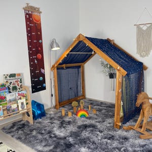 May include: A wooden playhouse with a navy blue starry canopy and a wooden rocking horse sits on a fluffy gray rug. A wooden bookshelf holds children's books. A solar system height chart hangs on the wall.